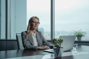 A confident businesswoman in a grey suit working on a laptop in a modern office setting.
